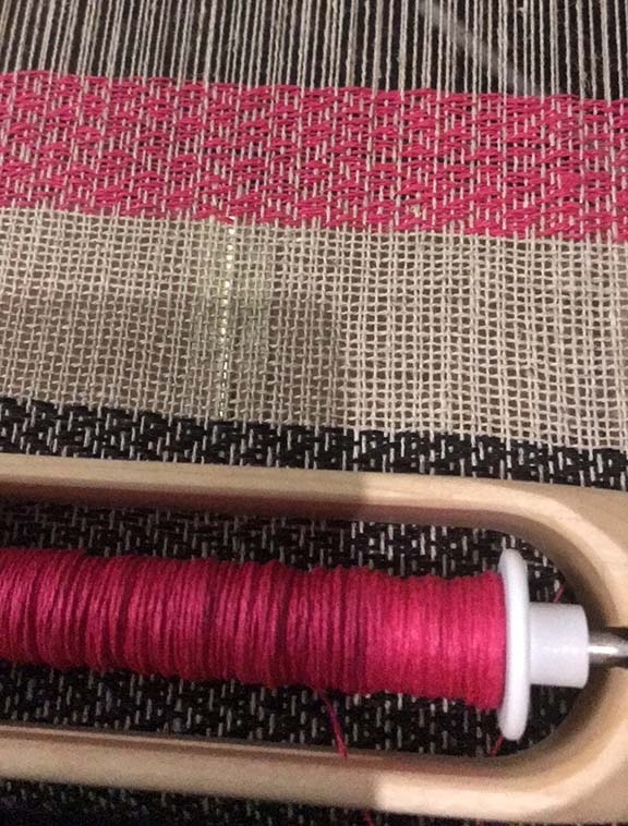 A close up of a couple of different weaves a vertical zig zag in black, a section of plain weave in off white and a more complex knot pattern in pink.