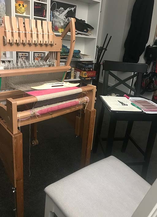 Image of my weaving workplace set up with the 8-shaft loom on a stand with a chair inform of it, next to the loom is a tall chair with a note book on it with my pattern notes and diagrams.