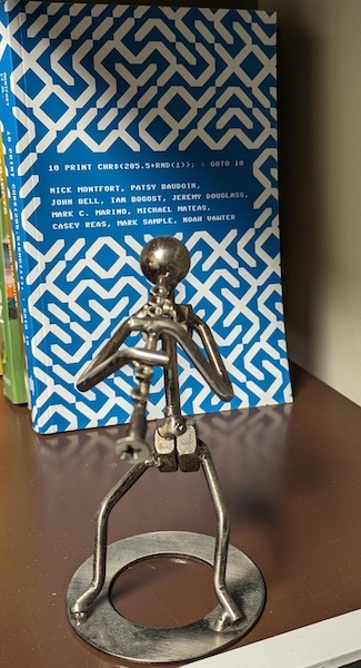 two photos of a metal figure in front of a book. The left shows the book "10 print chr" clearly while the right shows it blurred out.